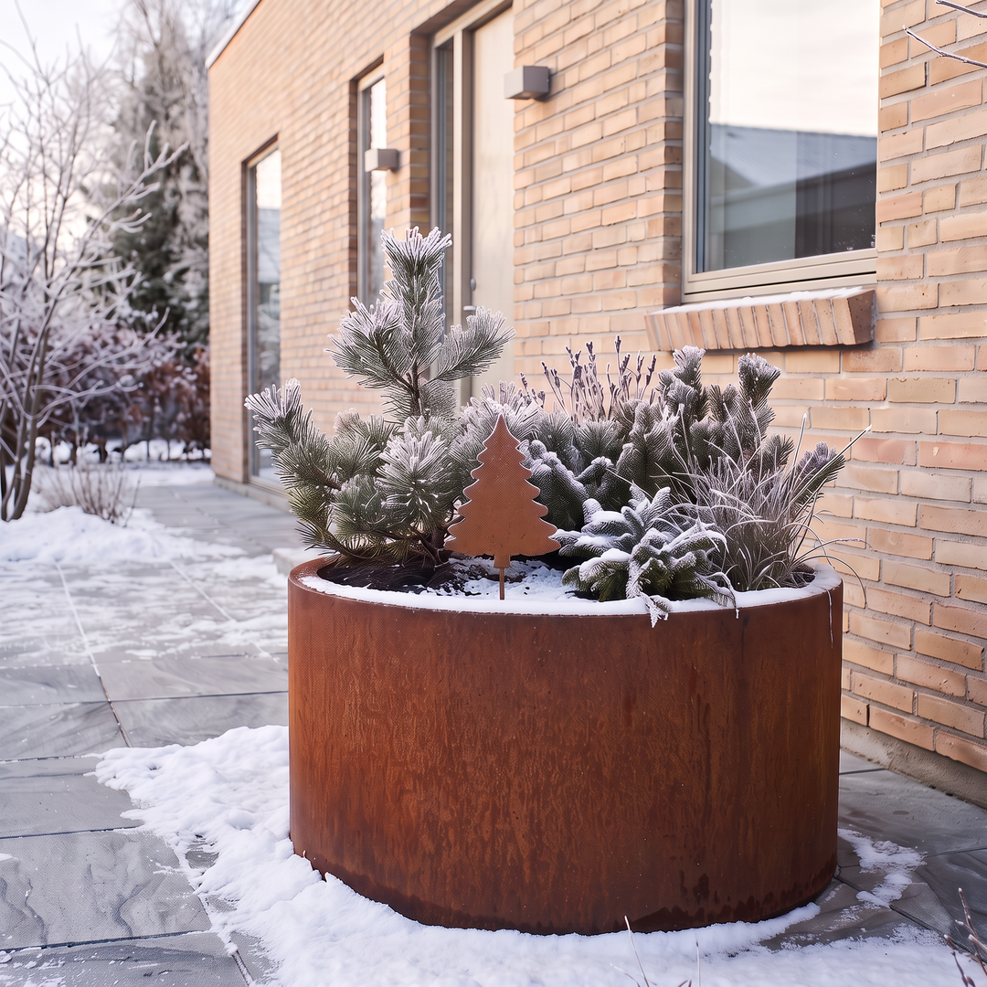 Gartenstecker in Tannenbaumform aus Cortenstahl in einem runden Pflanzkübel mit frostbedeckten Nadelgehölzen vor einer Backsteinfassade.