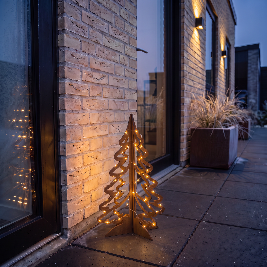 Weihnachtsbaum aus Cortenstahl mit leuchtender Lichterkette auf einer Terrasse an einer Backsteinwand am Abend.