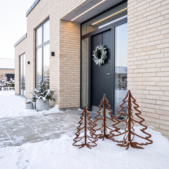 Drei Weihnachtsbäume aus Cortenstahl im Schnee vor einer dunklen Haustür mit Türkranz.