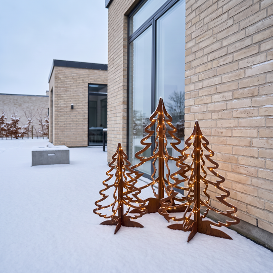 Beleuchtete Tannenbäume aus Cortenstahl in drei Größen auf einer verschneiten Terrasse direkt an der Hauswand.