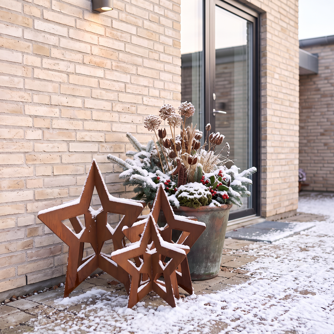 Rostbraune Deko-Sterne aus Cortenstahl mit Schnee auf gepflastertem Boden vor einer Haustür mit Glaseinsatz.