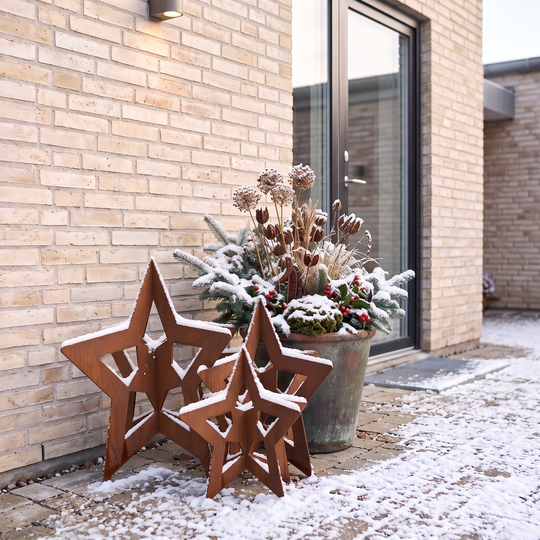 Rostbraune Deko-Sterne aus Cortenstahl mit Schnee auf gepflastertem Boden vor einer Haustür mit Glaseinsatz.